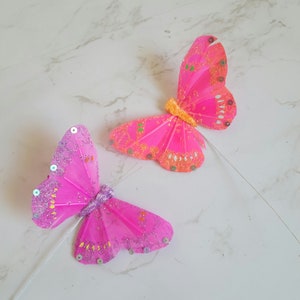 May include: Two pink butterfly decorations with glitter and sequins. The butterflies are attached to white stems.