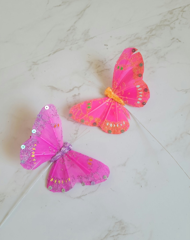 May include: Two pink butterfly decorations with glitter and sequins. The butterflies are attached to white stems.