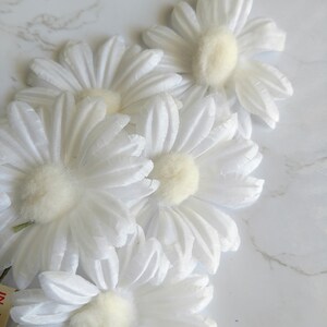 May include: Close-up of several white daisy flowers with soft, fluffy yellow centers. The petals are layered and textured, creating a delicate appearance. The flowers are arranged on a white marble surface.