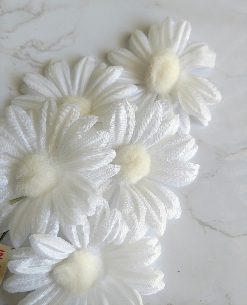May include: Close-up of several white daisy flowers with soft, fluffy yellow centers. The petals are layered and textured, creating a delicate appearance. The flowers are arranged on a white marble surface.