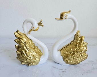 Puede incluir: Dos figuritas de cisne de cerámica blanca con detalles dorados. Los cisnes se miran con el cuello curvado. Las alas están detalladas con pan de oro.