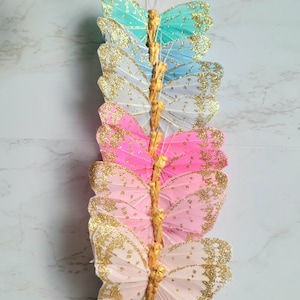 May include: A set of eight glittery butterfly decorations in various shades of blue, pink, and white. The butterflies are arranged vertically on a white background.