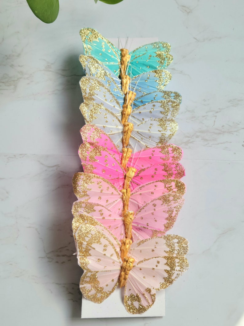 May include: A set of eight glittery butterfly decorations in various shades of blue, pink, and white. The butterflies are arranged vertically on a white background.