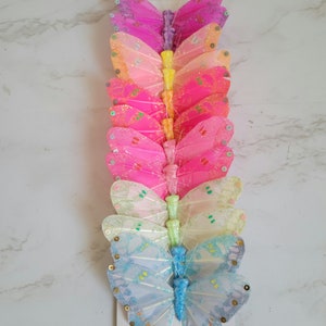 May include: A set of ten glittery butterfly embellishments in various pastel colors, including pink, blue, yellow, and green. The butterflies are arranged vertically in a stack, with the largest butterfly at the bottom and the smallest at the top.