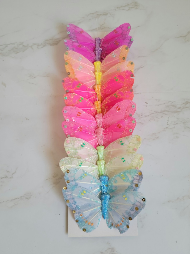 May include: A set of ten glittery butterfly embellishments in various pastel colors, including pink, blue, yellow, and green. The butterflies are arranged vertically in a stack, with the largest butterfly at the bottom and the smallest at the top.