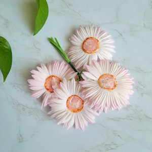 Puede incluir: Un ramo de cuatro margaritas artificiales de color rosa con centros naranjas sobre un fondo de m&aacute;rmol blanco.