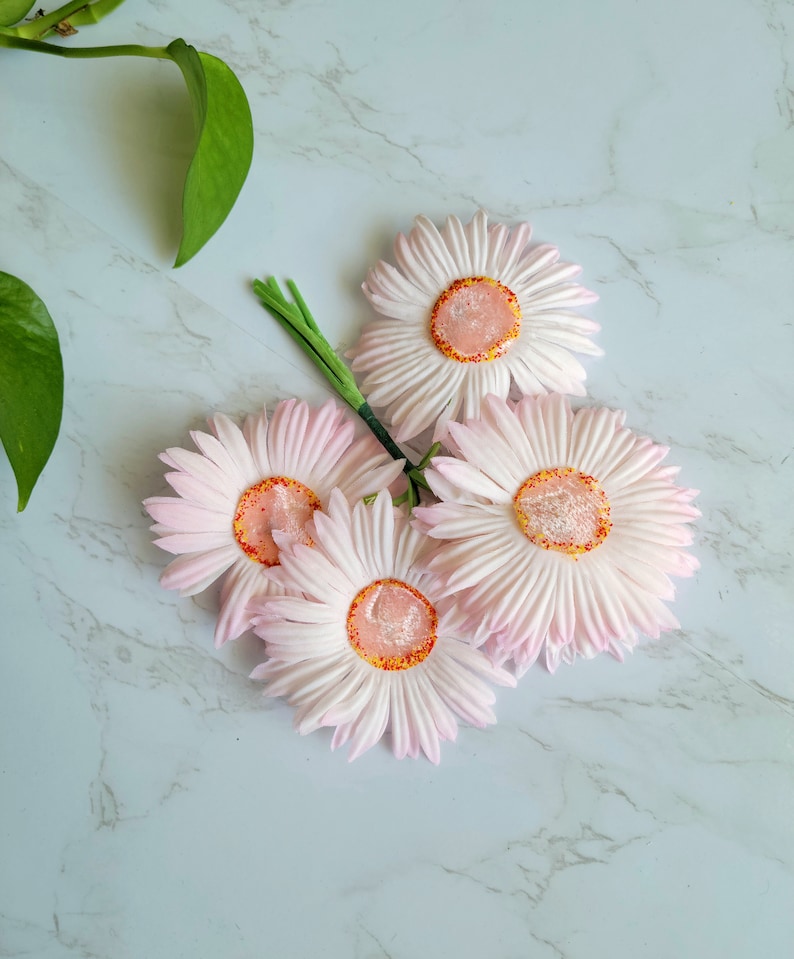 Puede incluir: Un ramo de cuatro margaritas artificiales de color rosa con centros naranjas sobre un fondo de m&aacute;rmol blanco.