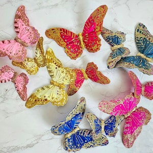 Puede incluir: Surtido de decoraciones de mariposas coloridas con detalles de purpurina. Las mariposas están hechas de papel y tienen una variedad de colores, incluyendo rosa, azul, rojo y amarillo.