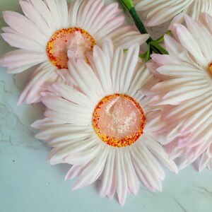Puede incluir: Un ramo de cuatro margaritas artificiales de color rosa y blanco con centros naranja y rojo. Las margaritas est&aacute;n dispuestas en un racimo y tienen una textura suave y aterciopelada.