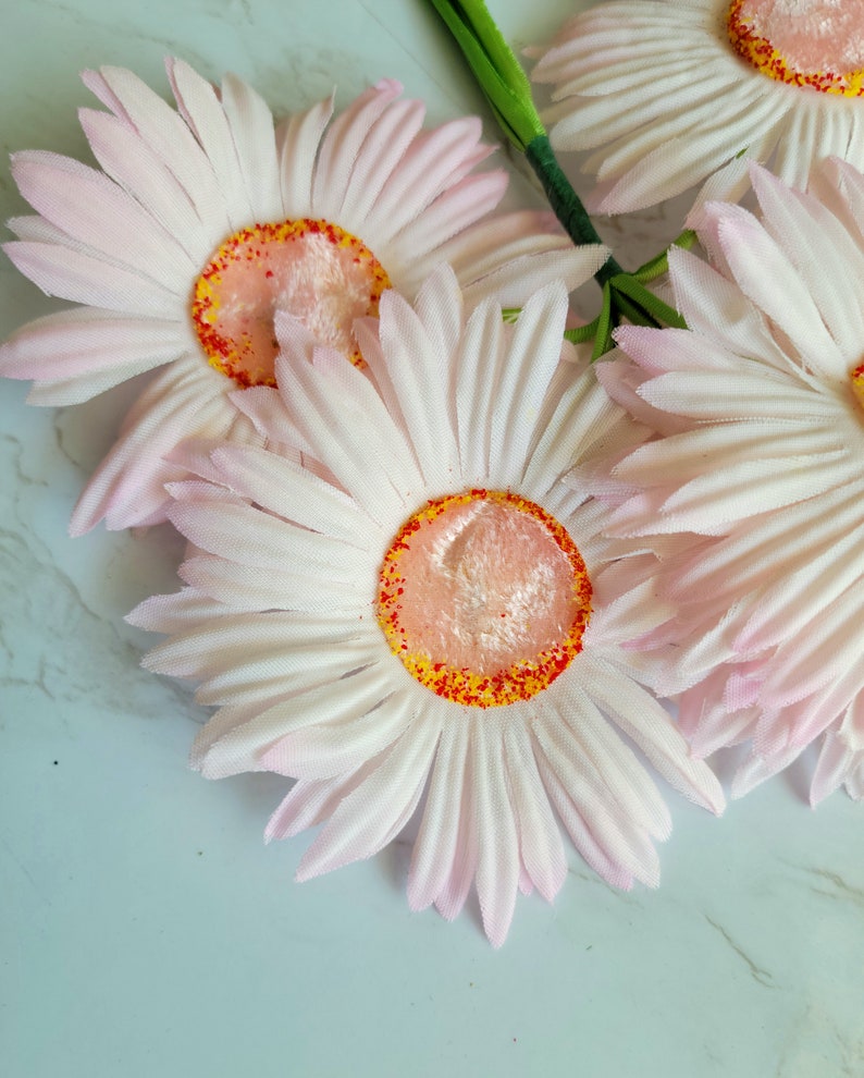 Puede incluir: Un ramo de cuatro margaritas artificiales de color rosa y blanco con centros naranja y rojo. Las margaritas est&aacute;n dispuestas en un racimo y tienen una textura suave y aterciopelada.