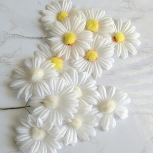 May include: A collection of white daisy flower stems with yellow centers and red accents. The petals are layered and textured. The flowers are arranged on a white marble surface.
