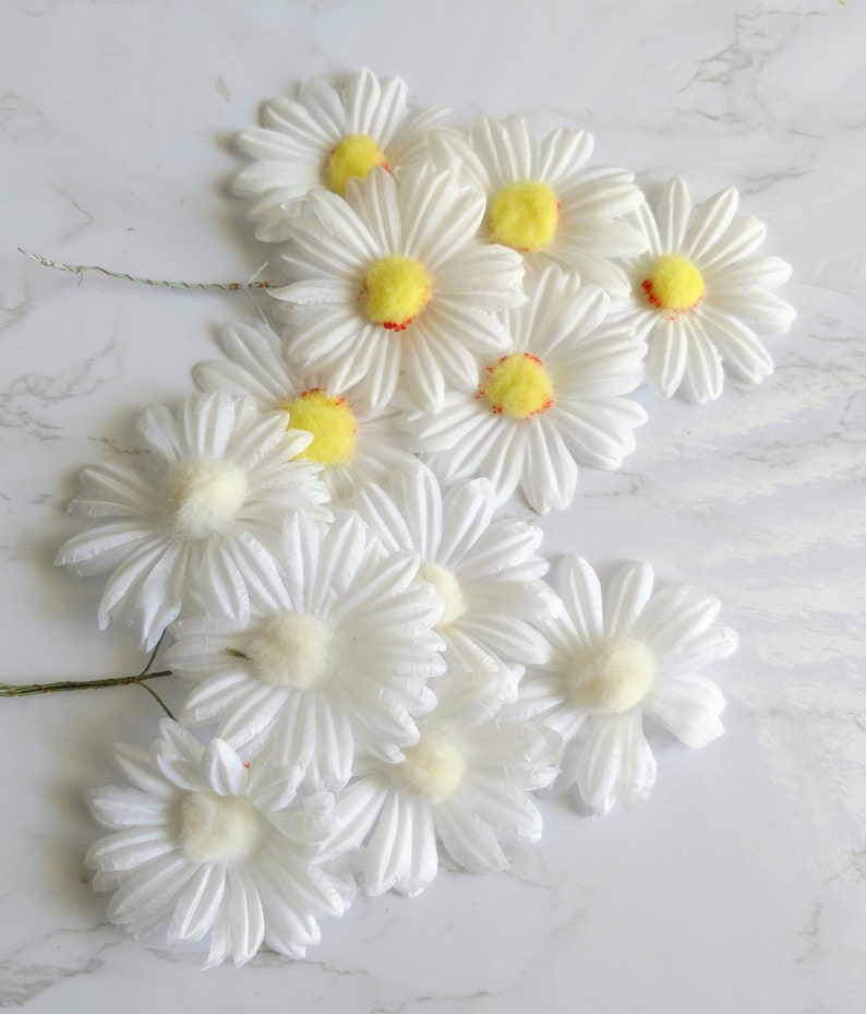 May include: A collection of white daisy flower stems with yellow centers and red accents. The petals are layered and textured. The flowers are arranged on a white marble surface.