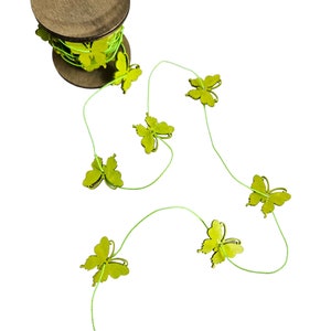 May include: A wooden spool with green string and attached lime green butterfly decorations. Several butterflies are strung along the green thread, creating a decorative garland. The spool is brown and the butterflies are a bright, cheerful green.
