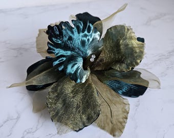 Peut inclure: Une grande fleur décorative avec des pétales superposés dans les tons de bleu sarcelle, vert olive et beige. Le centre de la fleur présente un motif texturé bleu sarcelle et les pétales sont ornés de petits embellissements scintillants. Il s'agit probablement d'un accessoire pour cheveux.