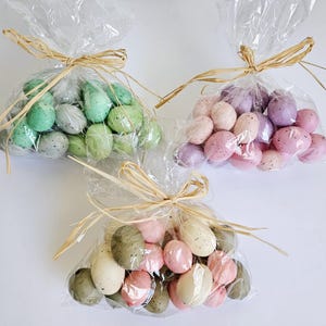 May include: Three bags of pastel coloured speckled Easter eggs. The bags are tied with twine and are filled with green, pink and purple eggs.