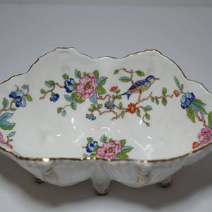 May include: A white porcelain serving dish with a scalloped edge and three feet. The dish is decorated with a floral pattern in shades of pink, blue, and green, and features a small bird in flight.