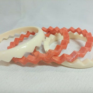 May include: Set of four plastic bangles, two are white and two are coral pink. The bangles have a zig-zag pattern around the edge.