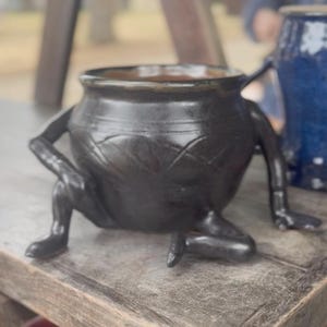 May include: A dark brown ceramic pot with a rounded body and a wide opening. The pot features three sculpted legs and one arm extending from the side. The exterior surface has a glossy finish with a subtle cross-hatched pattern etched into the clay.