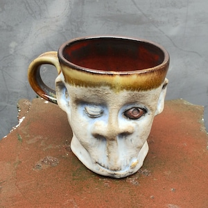 May include: A ceramic mug shaped like a face with a brown and yellow glaze. The mug has a single eye and a wide, open mouth.