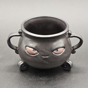 May include: A black ceramic cauldron with two handles and a grumpy face. The cauldron has two small feet and brown eyes.
