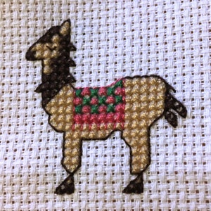 May include: A brown and tan llama with a red, green, and white saddle. The llama is embroidered on a white fabric.