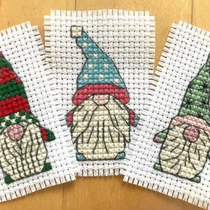 May include: Three cross-stitch ornaments featuring gnomes with different colored hats. The gnome on the left has a red and green hat, the middle gnome has a blue and white hat, and the gnome on the right has a green hat.