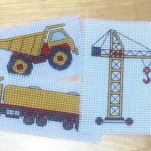 May include: Three cross-stitch patterns featuring construction vehicles: a yellow dump truck, a yellow cement mixer truck, and a yellow crane.