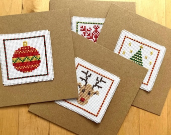 May include: A collection of handmade Christmas cards with cross-stitched designs. Each card features a different festive motif: a red ornament, a snowflake, a Christmas tree, and a reindeer. The cards are on brown cardstock.