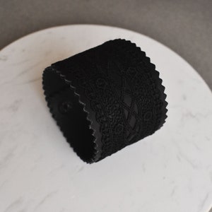 以下が含まれることがあります： 繊細なレースのデザインが施された黒い革製のカフブレスレットです。