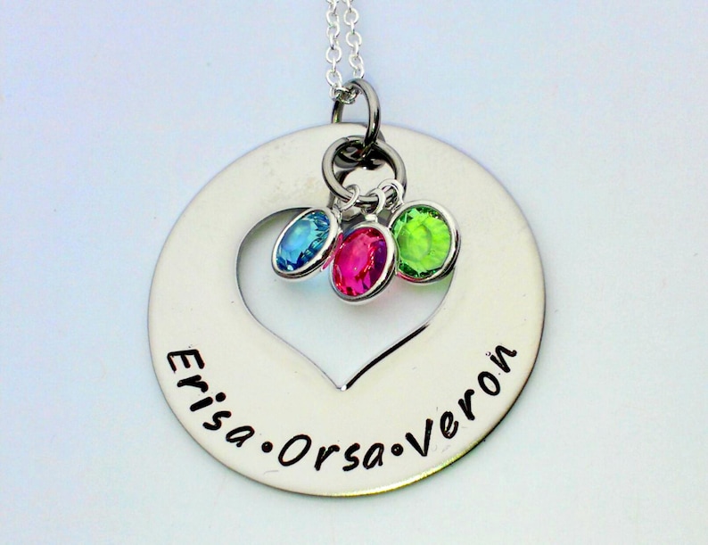 K&ouml;nnte beinhalten: Silberkette mit einem runden Anh&auml;nger, der ein ausgeschnittenes Herz und die eingravierten Namen "Errisa", "Orsa" und "Veron" zeigt. Drei kleine Edelsteine, einer blau, einer rosa und einer gr&uuml;n, sind am Anh&auml;nger befestigt.