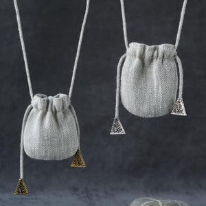 Puede incluir: Dos pequeñas bolsas grises con cordón y detalles triangulares decorativos de metal cuelgan de cuerdas blancas. Las bolsas están hechas de una tela texturizada. Debajo hay una colección de piedras, incluyendo pirita y una roca redondeada.