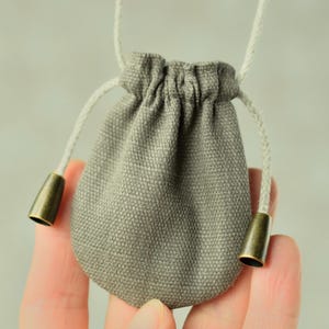 Könnte beinhalten: Ein kleiner, grauer Beutel mit Kordelzug, weißer Kordel und bronzefarbenen Metallakzenten. Der Beutel wird in einer Hand gehalten und zeigt seine kompakte Größe und sein schlichtes Design. Der Beutel besteht aus einem strukturierten Stoff.
