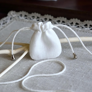 May include: A small, white, woven fabric drawstring pouch featuring a simple closure mechanism with two long white cords. Each cord end is finished with a polished, silver-toned metal bead, providing a clean and functional design for this accessory.