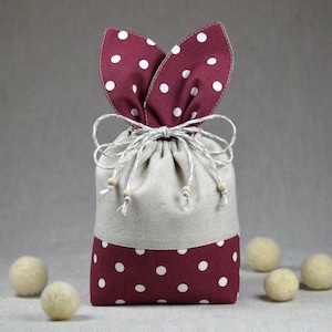 Może przedstawiać: Mała, beżowa torba ze sznurkiem do ściągania z uszami króliczymi z czerwono-białej tkaniny w kropki i dnem z czerwono-białej tkaniny w kropki. Torba jest związana naturalnym sznurkiem.