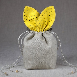 Puede incluir: Una pequeña bolsa de cordón de lino beige con orejas de conejo de tela amarilla con lunares negros. La bolsa tiene dos cordones largos de color beige con cuentas de madera en los extremos.