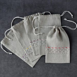 Puede incluir: Una colección de cinco bolsas grises de lino con cordón. Cada bolsa presenta un ribete de encaje blanco con sutiles acentos de color amarillo, morado y rojo. Las bolsas están dispuestas sobre una superficie gris oscuro, mostrando su diseño sencillo y textura natural.