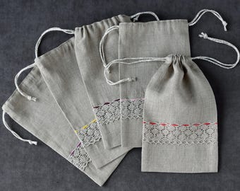 May include: A collection of five gray linen drawstring bags. Each bag features a white lace trim with subtle color accents of yellow, purple, and red. The bags are arranged on a dark gray surface, showcasing their simple design and natural texture.