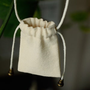 May include: A small, white drawstring bag with a white cord and brass toggles. The bag is hanging from a branch.