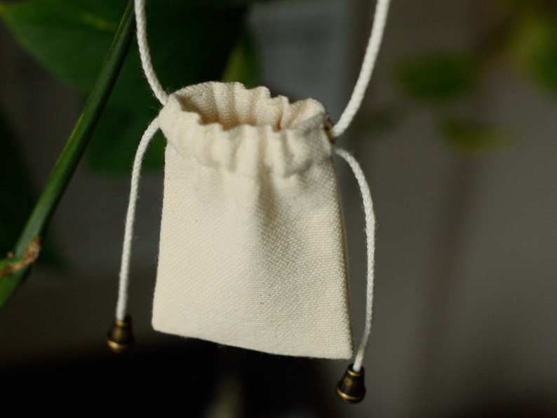 May include: A small, white drawstring bag with a white cord and brass toggles. The bag is hanging from a branch.
