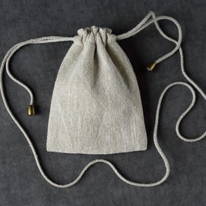 May include: A small, beige drawstring bag made of textured fabric. The bag has a rounded bottom and a gathered top, with a white cord that serves as a drawstring. The cord ends with small, gold-colored metal tips.