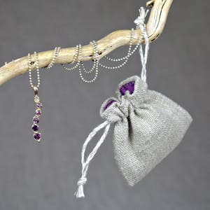 May include: A silver chain necklace with a purple gemstone pendant hangs from a branch. The necklace is displayed with a small, beige drawstring bag.