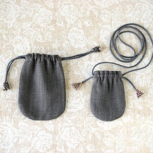 May include: Two gray drawstring pouches with decorative metal accents. The pouches are made of a textured fabric and have a rounded bottom. The drawstring is a matching gray cord.
