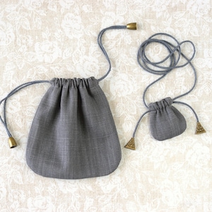 May include: Two grey drawstring pouches of different sizes. The larger pouch has a rounded bottom and a drawstring closure. The smaller pouch has a similar design. Both have grey drawstrings with gold-coloured metal accents.