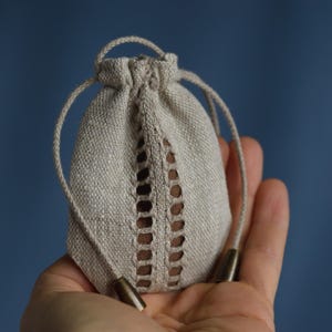 Puede incluir: Una pequeña bolsa beige con cordón y un diseño calado decorativo en la parte delantera. La bolsa tiene un aspecto texturizado y está hecha de una tela de aspecto natural. El cordón es un cordón beige claro con puntas metálicas.