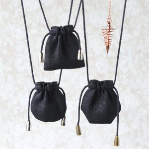 Könnte beinhalten: Drei kleine schwarze Beutel mit Kordelzug und metallischen Akzenten hängen an schwarzen Kordeln. Ein kupferfarbenes Pendel mit Spiralmuster hängt an einer Kette. Die Beutel sind aus einem strukturierten Stoff.