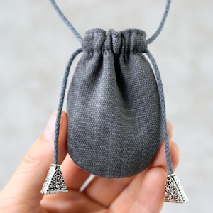 Op de afbeelding: Een kleine, grijze zak met trekkoord van een geweven stof. De zak heeft een gerimpelde bovenkant en wordt opgehangen aan een grijs koord. Het koord eindigt met decoratieve zilverkleurige metalen kegelvormige accenten. De zak wordt in een hand gehouden.