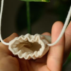 May include: A small, white drawstring pouch with a gathered top, hanging from a white cord. The pouch is held open by a hand, revealing the empty interior.