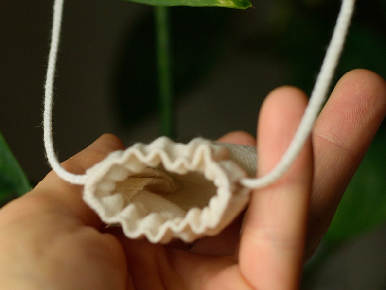 May include: A small, white drawstring pouch with a gathered top, hanging from a white cord. The pouch is held open by a hand, revealing the empty interior.