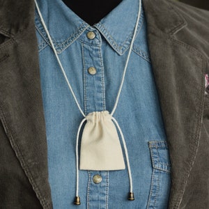 May include: A white cotton drawstring pouch hanging from a white cord around a blue denim shirt collar. The pouch is hanging over a brown corduroy jacket.