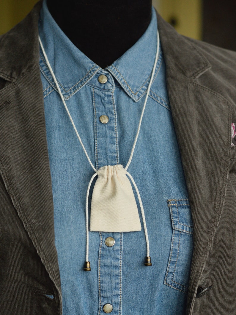 May include: A white cotton drawstring pouch hanging from a white cord around a blue denim shirt collar. The pouch is hanging over a brown corduroy jacket.